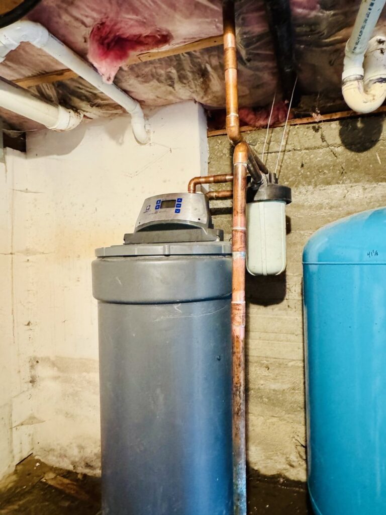 A water softener system and pressure tank in a basement with visible water stains on the floor and wall, indicating water damage, by The Plumbing Dr in Falls Church, VA