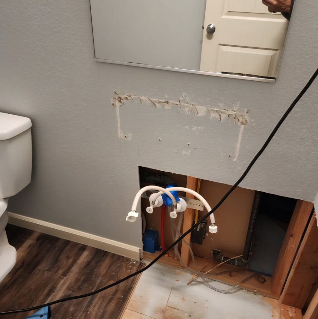 A bathroom wall with a section cut out, revealing pipes and subfloor damage for water damage repair by Verg Restoration in Vancouver, WA