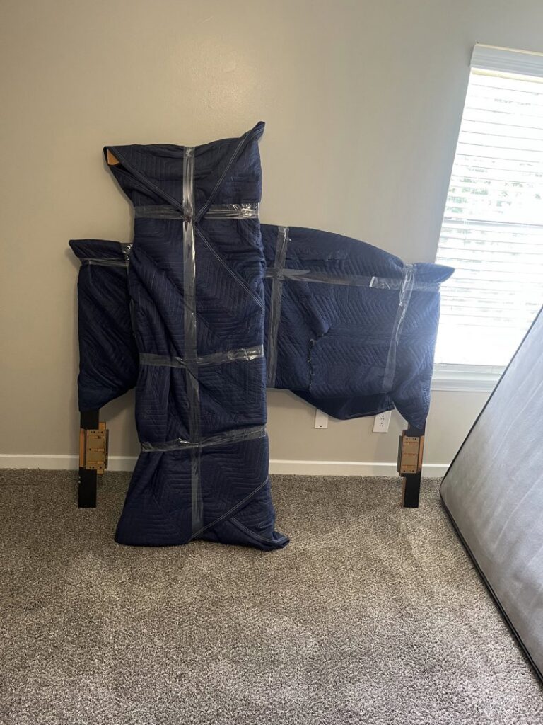 A bed frame and headboard carefully wrapped in moving blankets and tape for transport by Gifted Journey Movers in Gulfport, MS.