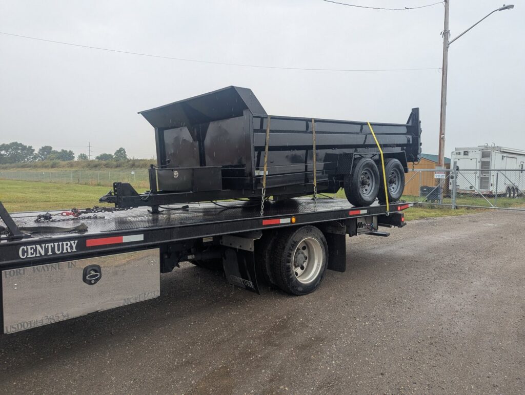 A black dump trailer being transported on a flatbed tow truck by First Hand Towing & Recovery in Fort Wayne, IN.