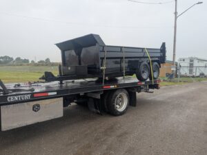 A black dump trailer being transported on a flatbed tow truck by First Hand Towing & Recovery in Fort Wayne, IN.