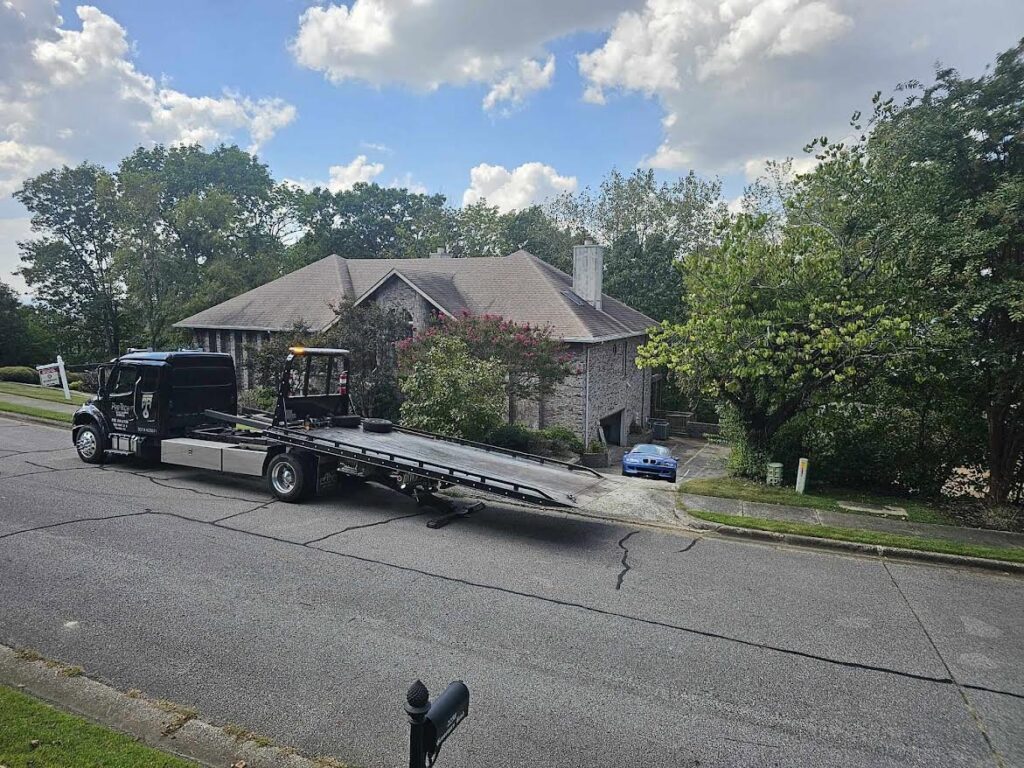 A black flatbed tow truck from Pro-Tech Wrecker Service preparing to load a blue car from a driveway in Decatur, AL.