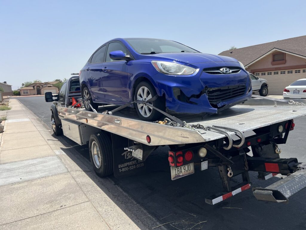 A black flatbed tow truck from Poly's Towing Glendale transporting a damaged blue Hyundai sedan in Glendale, AZ.
