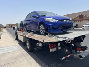 A black flatbed tow truck from Poly's Towing Glendale transporting a damaged blue Hyundai sedan in Glendale, AZ.