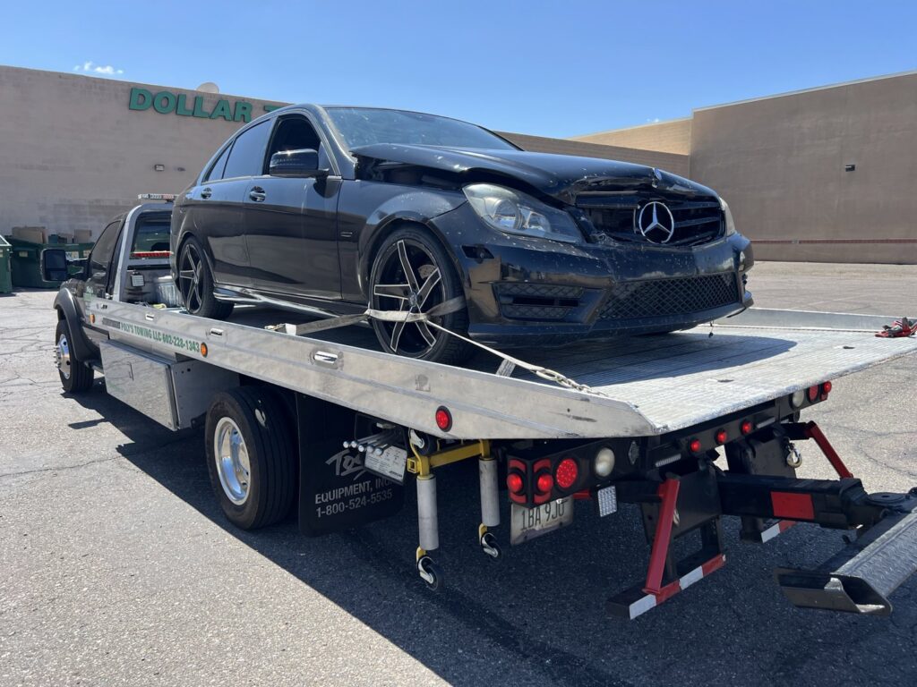 A black flatbed tow truck from Poly's Towing Glendale transporting a damaged black Mercedes-Benz sedan in Glendale, AZ.
