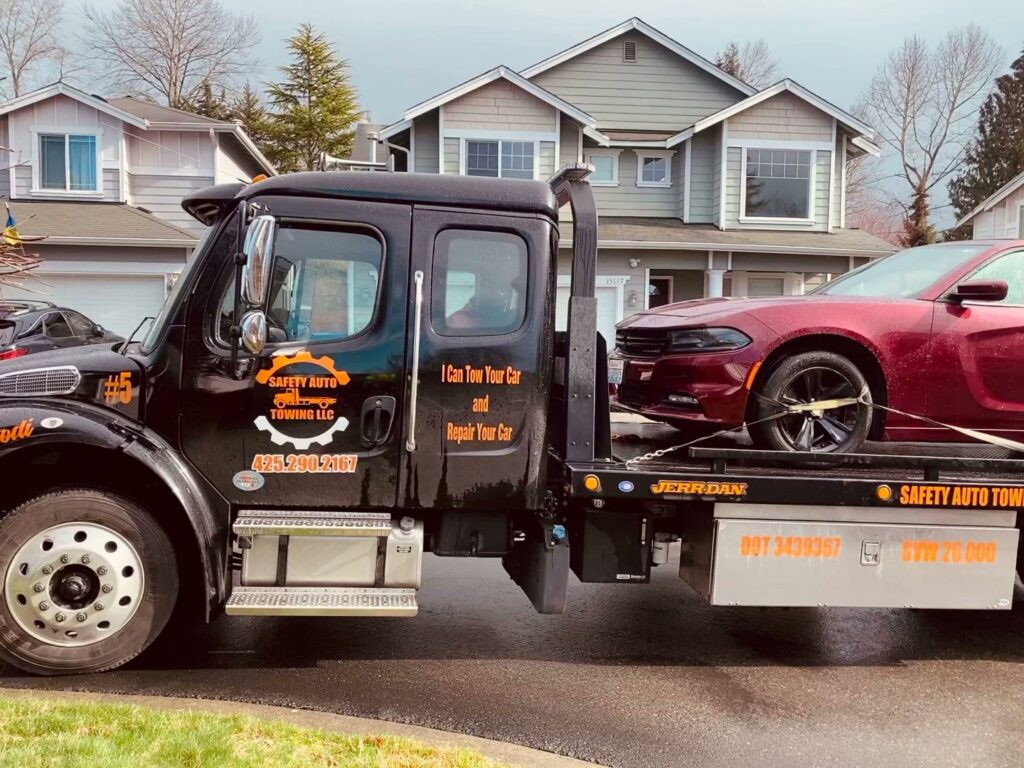 A black flatbed tow truck from Safety AUTO Towing LLC in Seattle, WA, transporting a red Dodge Charger.