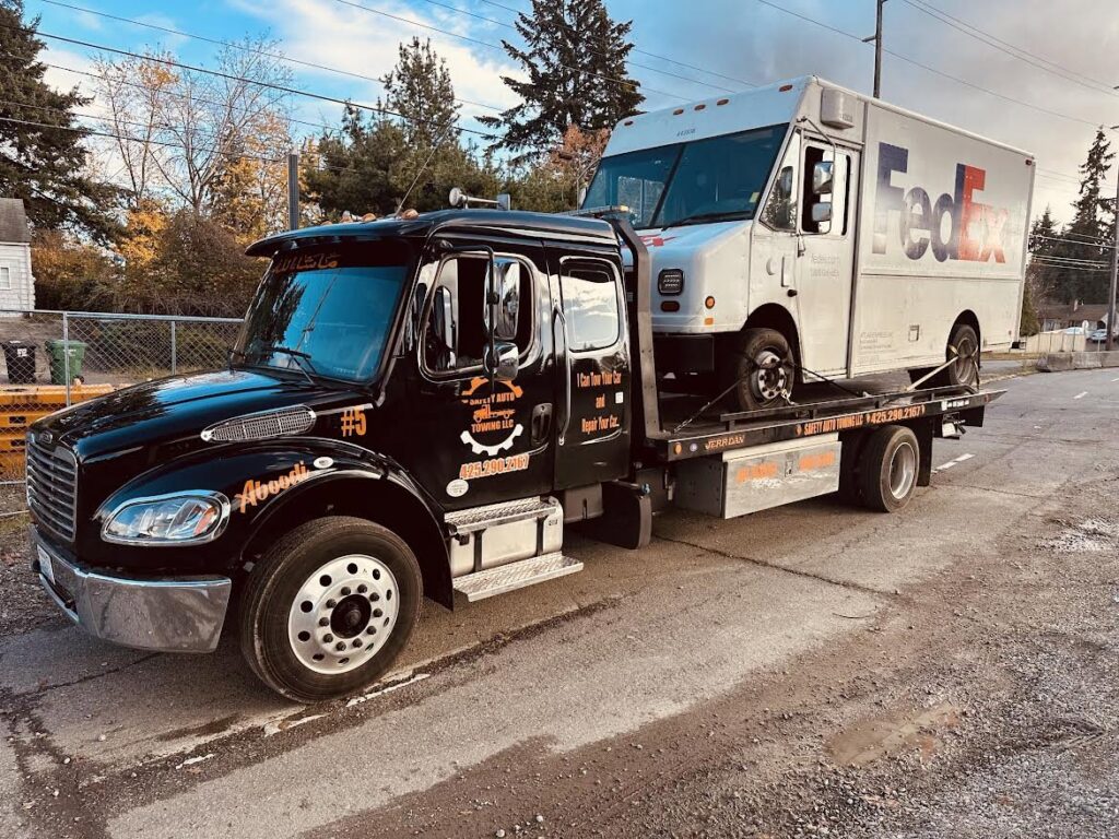 A black flatbed tow truck from Safety AUTO Towing LLC in Seattle, WA, transporting a FedEx delivery truck.