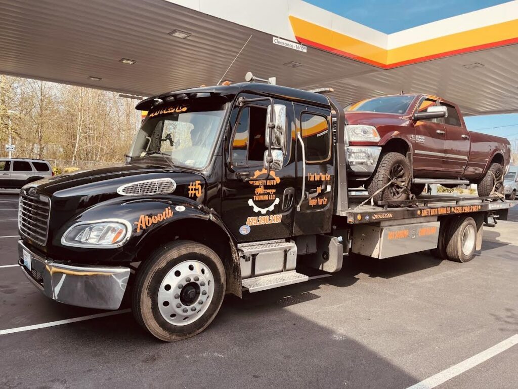 A black flatbed tow truck from Safety AUTO Towing LLC in Seattle, WA, transporting a red pickup truck at a gas station.