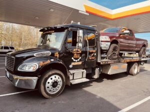 A black flatbed tow truck from Safety AUTO Towing LLC in Seattle, WA, transporting a red pickup truck at a gas station.