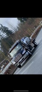 A black flatbed tow truck from Safety AUTO Towing LLC in Seattle, WA, transporting a silver car on a road.