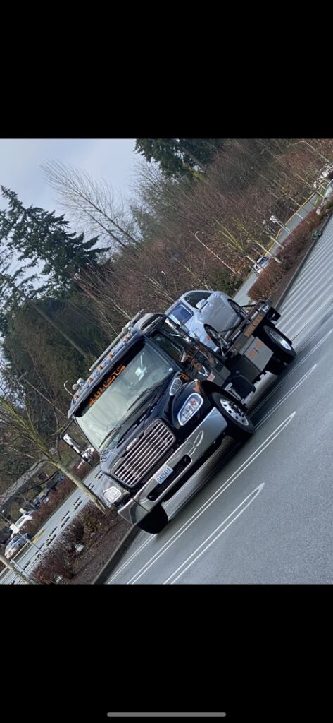 A black flatbed tow truck from Safety AUTO Towing LLC in Seattle, WA, transporting a silver car on a road.