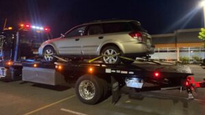 A black flatbed tow truck from Safety AUTO Towing LLC in Seattle, WA, transporting a silver station wagon at night.