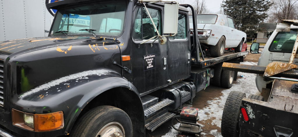 A black flatbed tow truck transporting an old white car for Boyz Automotive & Towing in Toledo, OH.