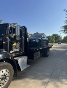 A black flatbed tow truck from BUVO towing services transporting a black car on a residential street in Fort Worth, TX.