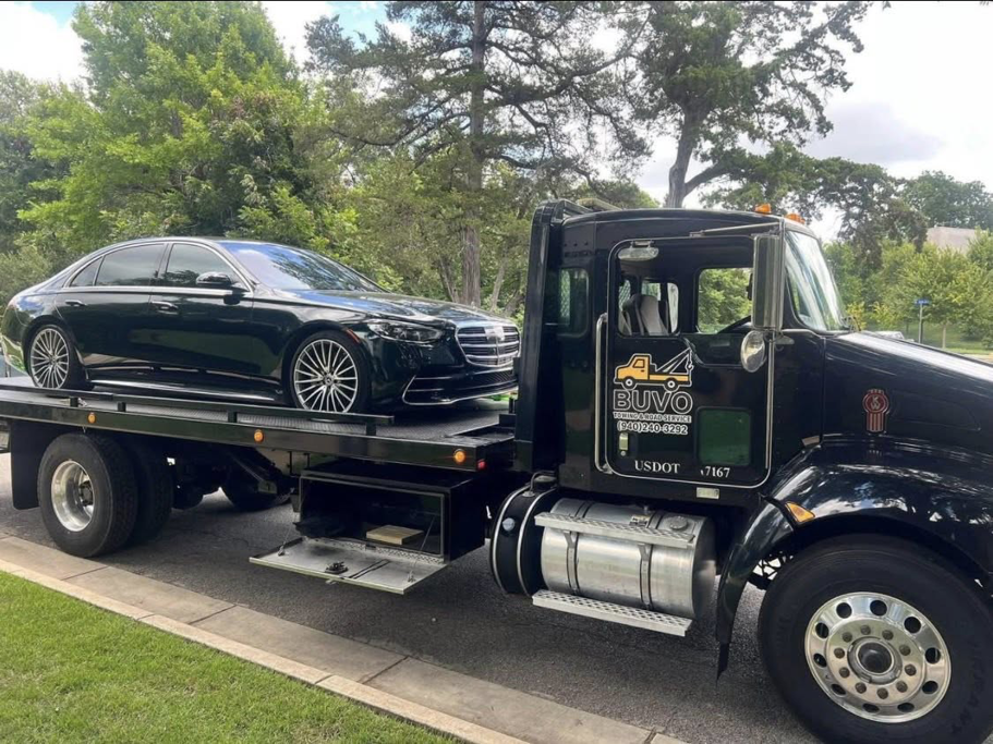 A black flatbed tow truck from BUVO towing services transporting a luxury black sedan on a street in Fort Worth, TX.