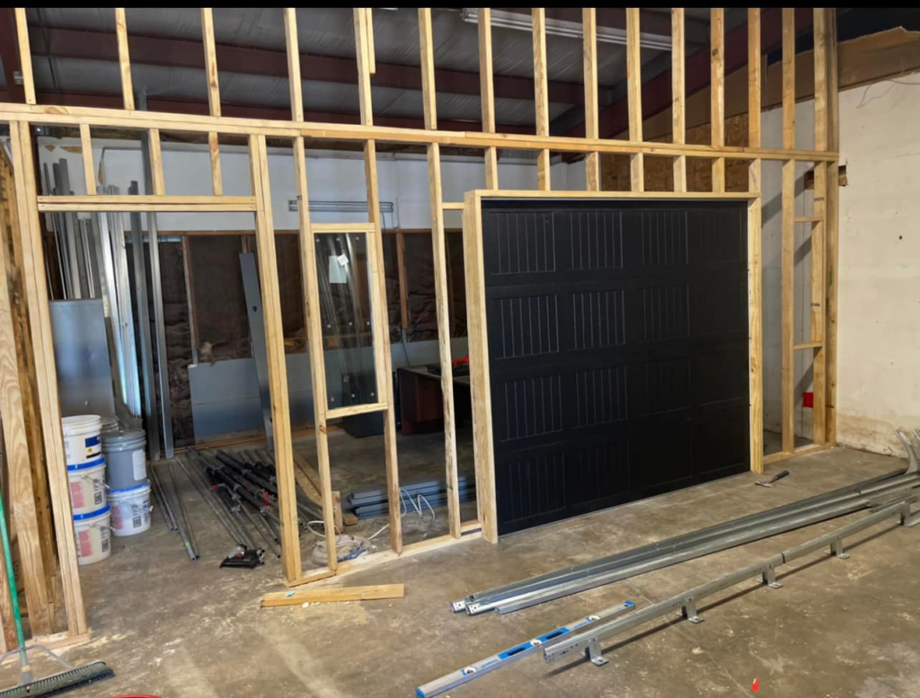 A black garage door being installed within a wooden framed wall, with tracks and components on the floor, by AMR Distribution Center in Wichita, KS