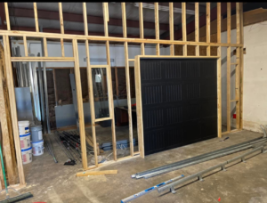 A black garage door being installed within a wooden framed wall, with tracks and components on the floor, by AMR Distribution Center in Wichita, KS