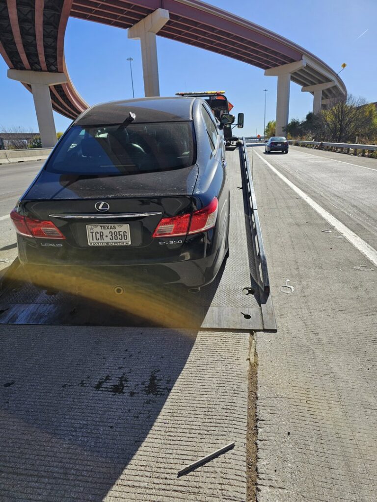 A black Lexus sedan being towed on a Tow Jam flatbed tow truck on a highway in Dallas, TX.
