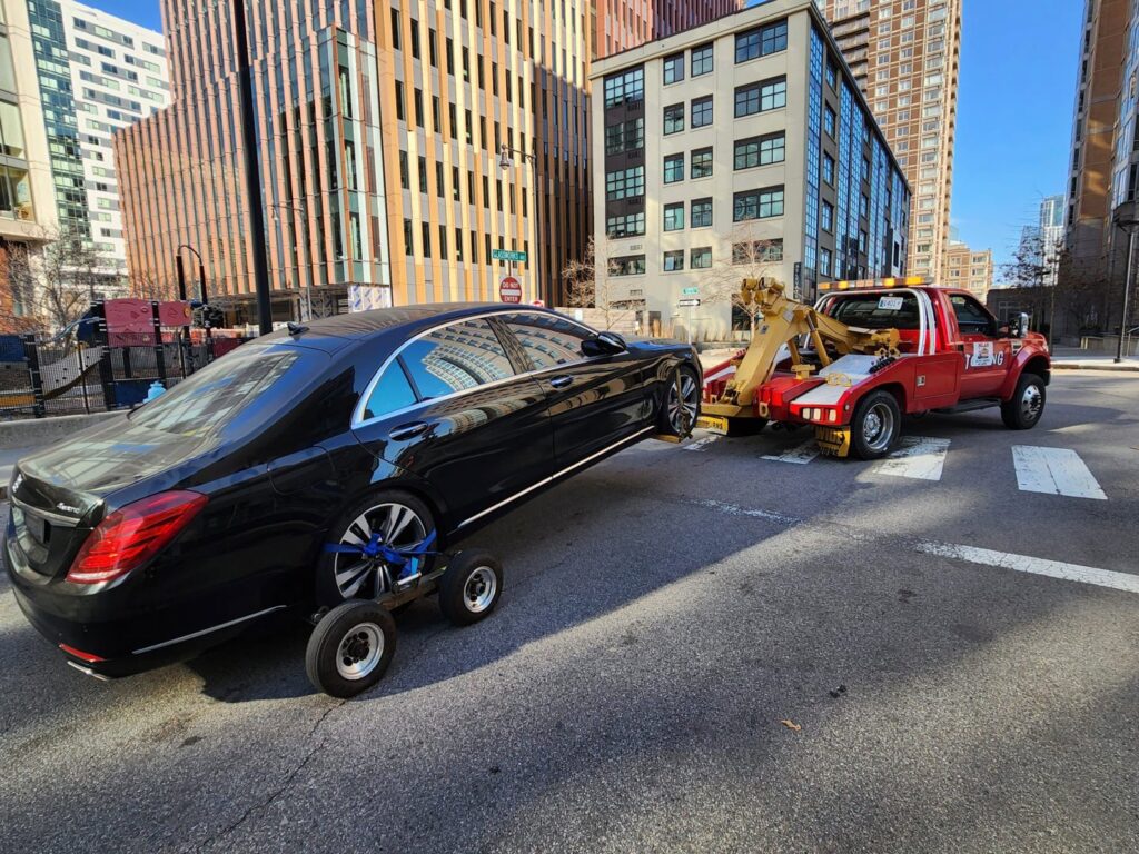 A black luxury sedan being towed by a red wheel-lift tow truck with dollies by Milan Towing inc. in Milan, MI.