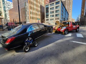 A black luxury sedan being towed by a red wheel-lift tow truck with dollies by Milan Towing inc. in Milan, MI.