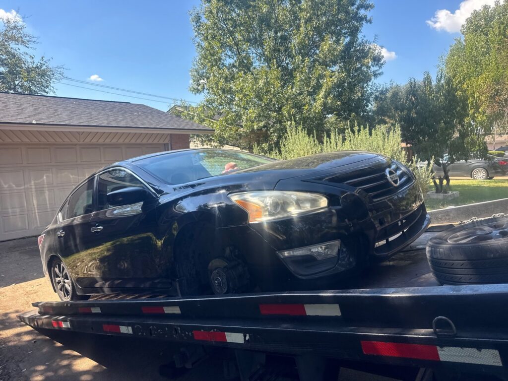 A black Nissan sedan with a missing front wheel being transported on a flatbed tow truck by 247 Towing in San Antonio, TX.