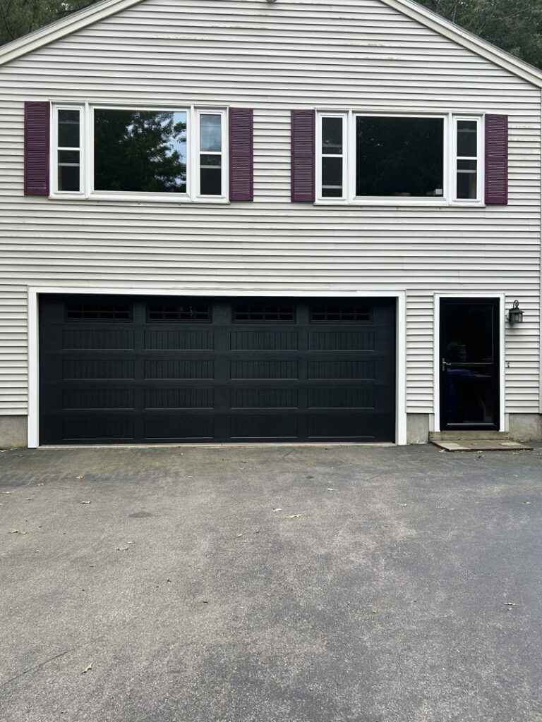 A black panel garage door with multiple windows installed on a light-colored house by NDI Garage Door in Plaistow, NH.