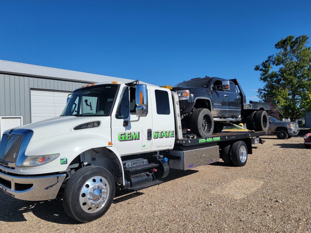 A large black pickup truck being transported on a flatbed tow truck by Gem State Towing and Recovery in Caldwell, ID.