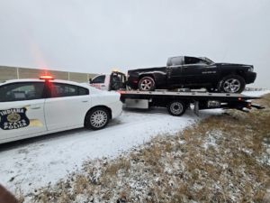 A black pickup truck loaded onto a Montes Towing Services flatbed truck, with an Indiana State Police car nearby, on a snowy road in Indianapolis, IN.