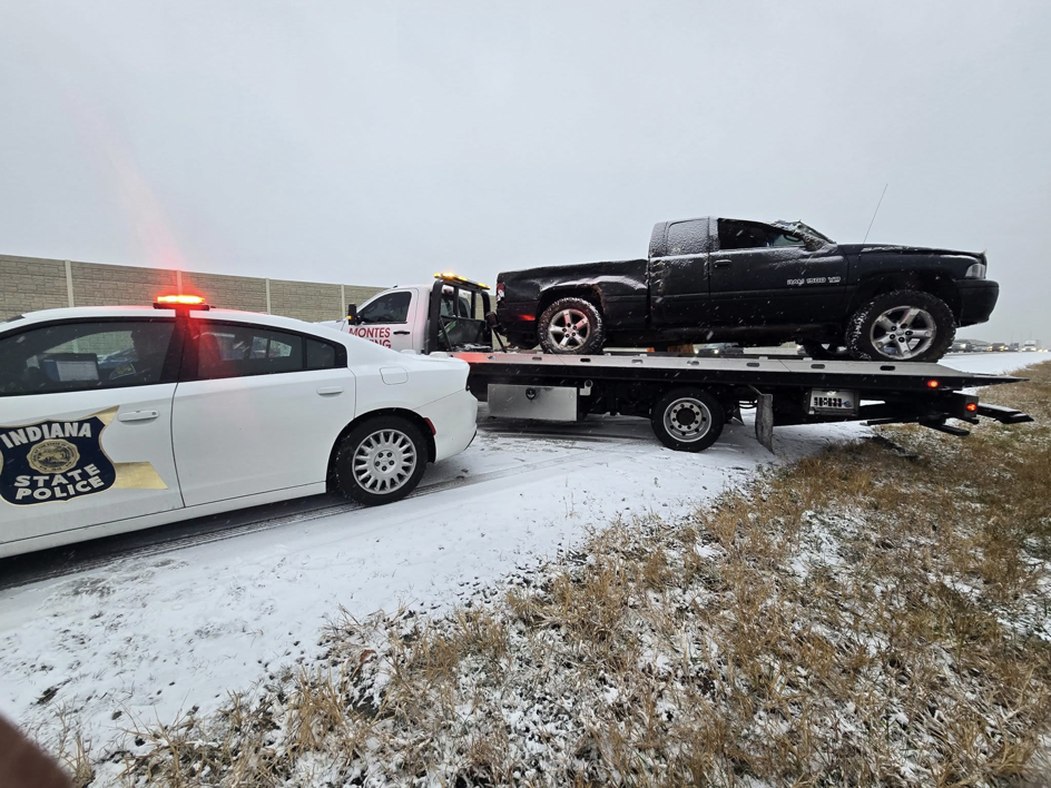A black pickup truck loaded onto a Montes Towing Services flatbed truck, with an Indiana State Police car nearby, on a snowy road in Indianapolis, IN.
