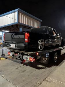 A black pickup truck being transported on a flatbed tow truck by Heavy Hookz Towing at night in Fort Worth, TX.