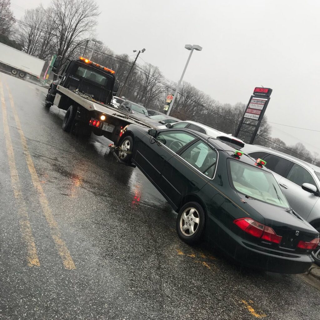 A black sedan being towed by a J & G Towing, LLC flatbed truck during rainy weather in Greensboro, NC.