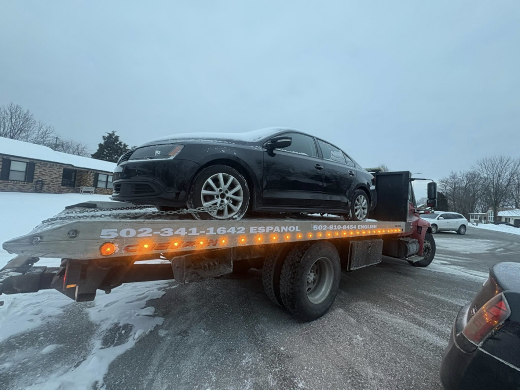 A black sedan covered in snow being towed on a flatbed truck by Tow & go in Louisville, KY.