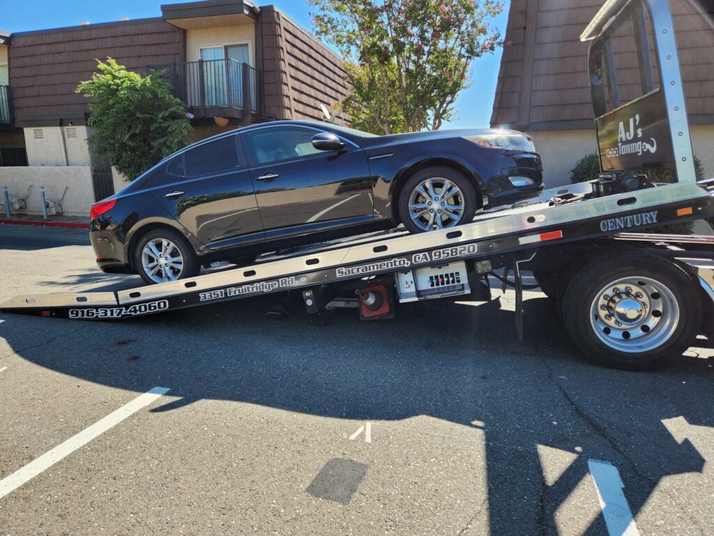 A black sedan being towed by an AJ's Towing Services & Roadside Assistance flatbed truck in Sacramento, CA.