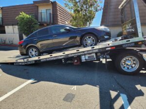 A black sedan being towed by an AJ's Towing Services & Roadside Assistance flatbed truck in Sacramento, CA.