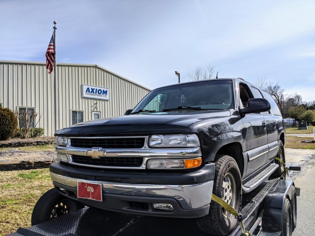 A black SUV being transported on a car trailer by Snoork Towing, parked in front of Axiom Foundations, LLC in Charlotte, NC.
