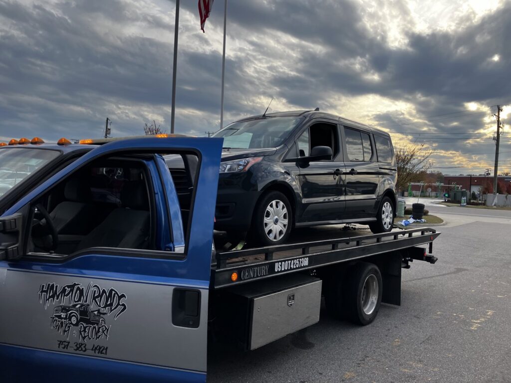 A black Tesla on a flatbed tow truck, towing a damaged blue sedan behind it by Hampton Roads Towing Services in Chesapeake, VA.