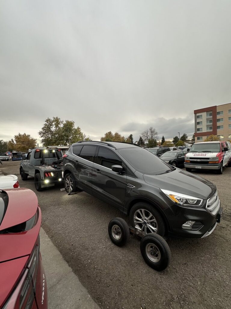 A black tow truck from Henderson Towing & Recovery LLC towing a black Tesla Model X in front of an apartment building in Denver, CO.