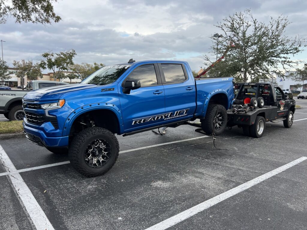 A black No Limit Towing truck actively towing a large blue pickup truck in a parking lot in Orlando, FL.