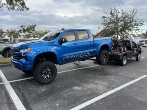 A black No Limit Towing truck actively towing a large blue pickup truck in a parking lot in Orlando, FL.