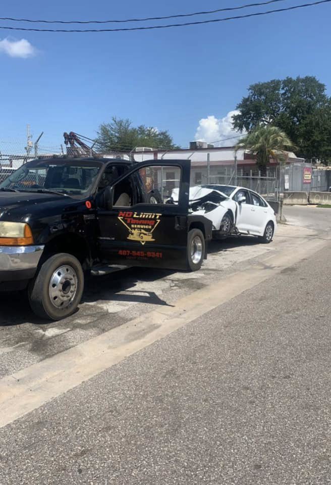 A black No Limit Towing truck actively towing a damaged white car on a sunny day in Orlando, FL.