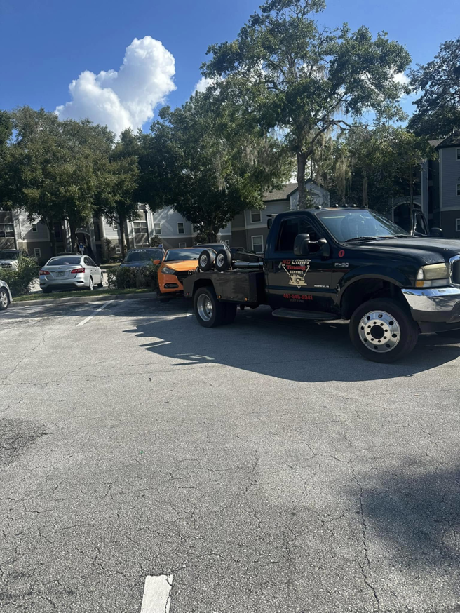 A black No Limit Towing truck actively towing an orange car in a residential parking lot in Orlando, FL.