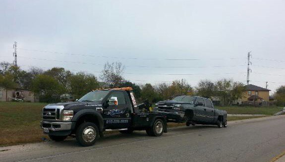 A black tow truck from Alanis Wrecker Service towing a black pickup truck on a road in San Antonio, TX.