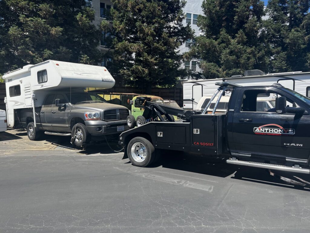 A black tow truck from Anthony's Towing & Transportation towing a large pickup truck with a camper shell in Yuba City, CA.