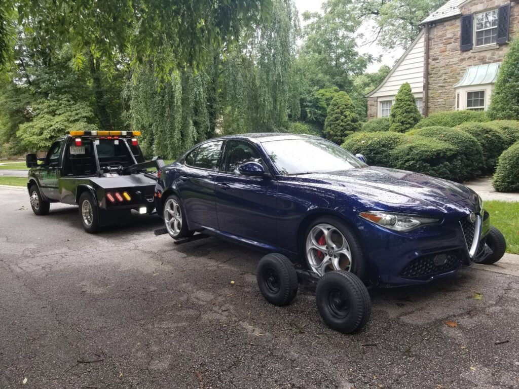 A blue Alfa Romeo sedan being towed by Whitelock Towing in Baltimore, MD, using a tow dolly.