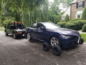 A blue Alfa Romeo sedan being towed by Whitelock Towing in Baltimore, MD, using a tow dolly.