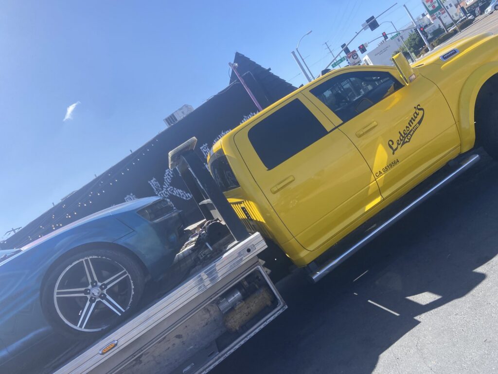 A blue car being transported on a yellow flatbed tow truck by Ledesma's Towing in San Diego, CA.