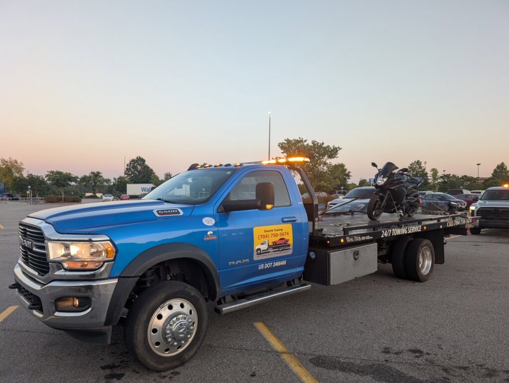 A blue flatbed tow truck from Snoork Towing transporting a black motorcycle in a parking lot in Charlotte, NC.