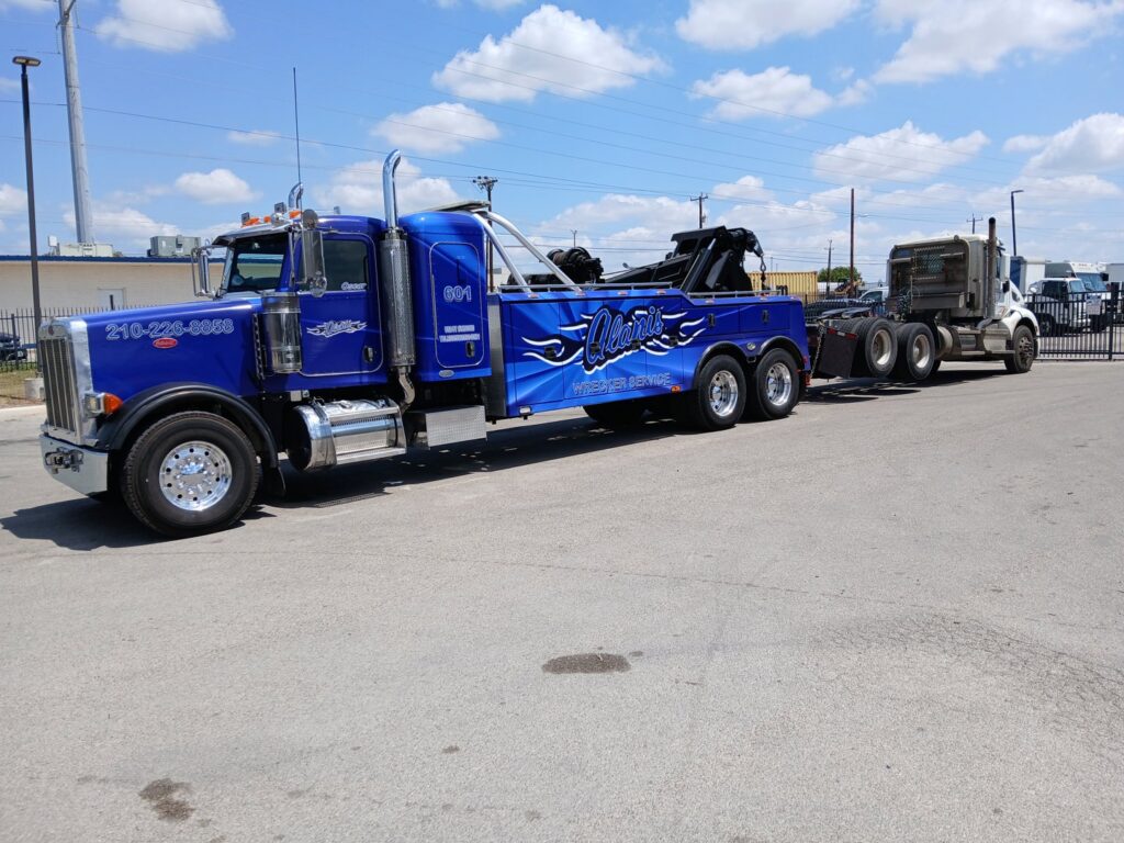 A blue heavy-duty tow truck from Alanis Wrecker Service towing a white semi-truck cab in San Antonio, TX.