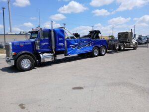 A blue heavy-duty tow truck from Alanis Wrecker Service towing a white semi-truck cab in San Antonio, TX.