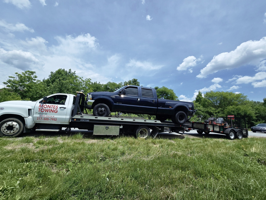Montes Towing Services flatbed truck towing a blue pickup truck and a small trailer on a sunny day in Indianapolis, IN.